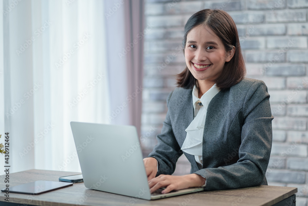 Beautiful young Asian business woman look at camera, using computer laptop technology. Browsing for online job while sitting at modern office desk. Professional manager secretary smile while working Beautiful young Asian business woman look at camera, using computer laptop technology. Browsing for online job while sitting at modern office desk. Professional manager secretary smile while working