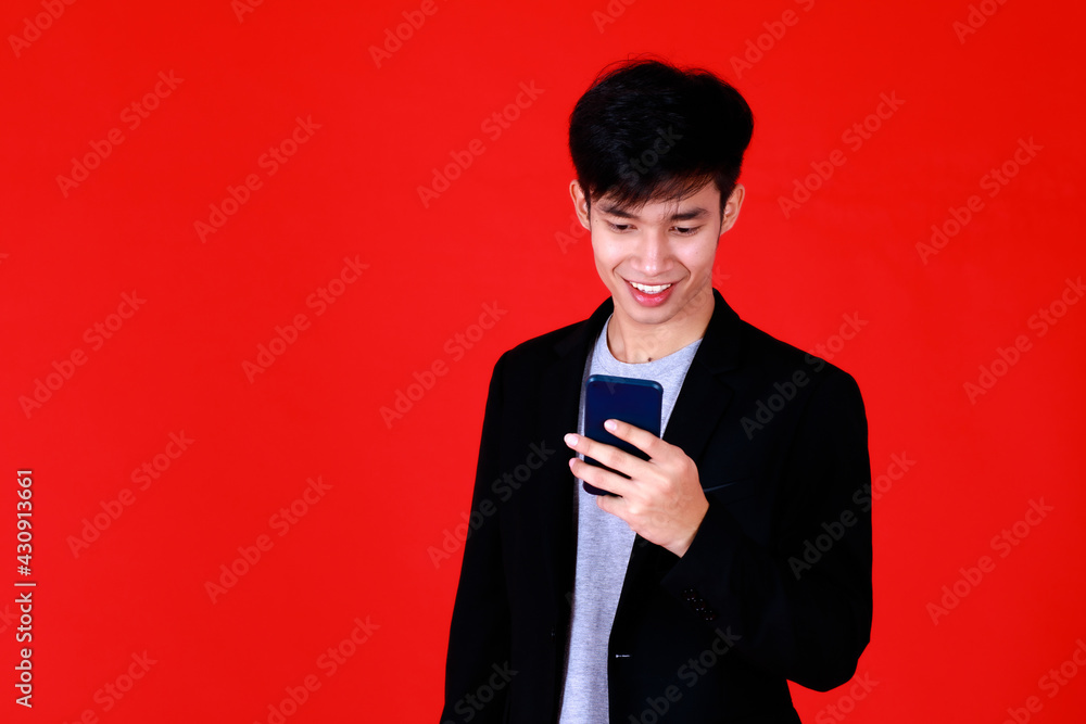 Asian young man standing look at smartphone screen smiling feel happy and confident. Shoot in studio with red background use to be design advertising or banner Asian young man standing look at smartphone screen smiling feel happy and confident. Shoot in studio with red background use to be design advertising or banner