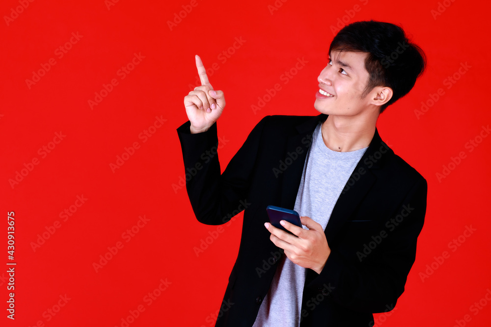 Asian young man standing look at camera smiling feel happy and confident.Hold smartphone and pointing to copy space.Shoot in studio with red background use to be design advertising or banner Asian young man standing look at camera smiling feel happy and confident.Hold smartphone and pointing to copy space.Shoot in studio with red background use to be design advertising or banner