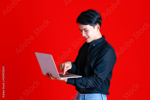 Asian young man standing look at camera and smile feel confident.Hold laptop on hand.Shoot in studio with red background with copy space use to be design advertising banner in concept tutor learning