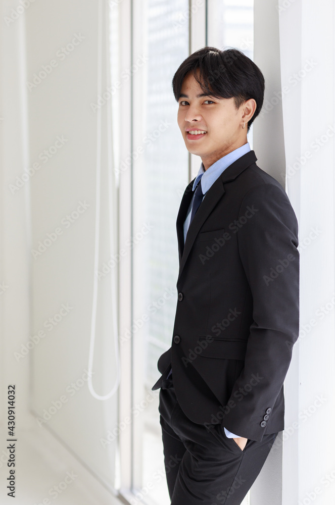 Portrait of young asian businessman who smart and handsome wearing black suit standing look at camera while smiling with look feel confident and window on background in office Portrait of young asian businessman who smart and handsome wearing black suit standing look at camera while smiling with look feel confident and window on background in office