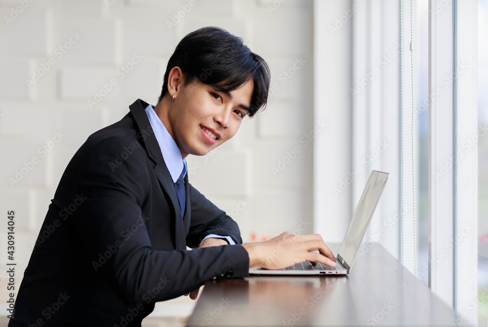 Asian young businessman are smart and handsome wearing black suit sitting while typing on keyboard at laptop near window in office at the morning Asian young businessman are smart and handsome wearing black suit sitting while typing on keyboard at laptop near window in office at the morning