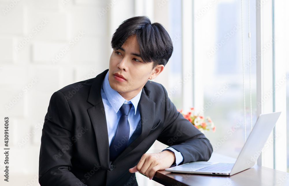 Asian young businessman are smart and handsome wearing black suit sitting while typing on keyboard at laptop near window in office at the morning Asian young businessman are smart and handsome wearing black suit sitting while typing on keyboard at laptop near window in office at the morning