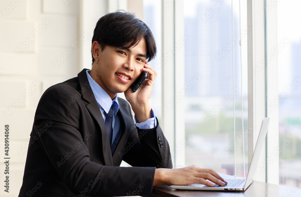 Portrait one young asian businessman are smart and handsome wearing black suit sitting chair talking phone while feel confident with smiling as smile on face looking at camera with laptop Portrait one young asian businessman are smart and handsome wearing black suit sitting chair talking phone while feel confident with smiling as smile on face looking at camera with laptop