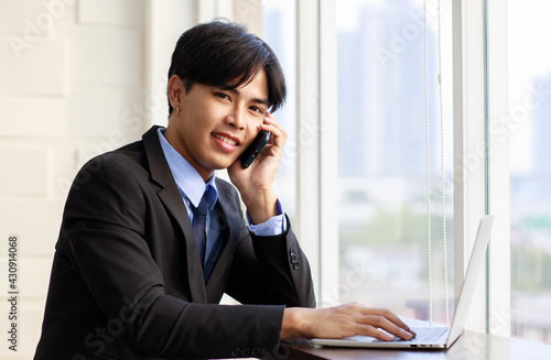 Portrait one young asian businessman are smart and handsome wearing black suit sitting chair talking phone while feel confident with smiling as smile on face looking at camera with laptop