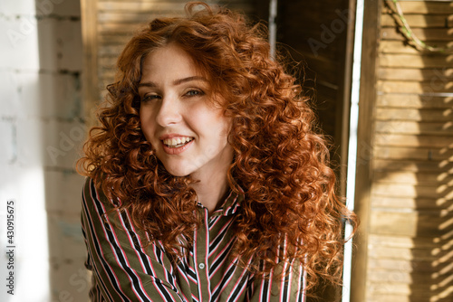 Portrait of happy redhead curly young woman near window smiling, close-up