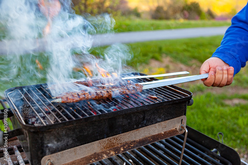 Cooking delicious Turkish Adana Kebab on a portable charcoal grill at a picnic location, Smoke and flame comes out of the sizzling meat. A man is turning the skewers around to prevent burning the meat