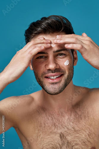 Cheerful good looking guy smiling at camera while applying white cream on his face, posing isolated over blue background