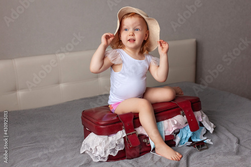 little girl surprised in beige summer hat sits on burgundy suitcase with clothes sticking out home on bed