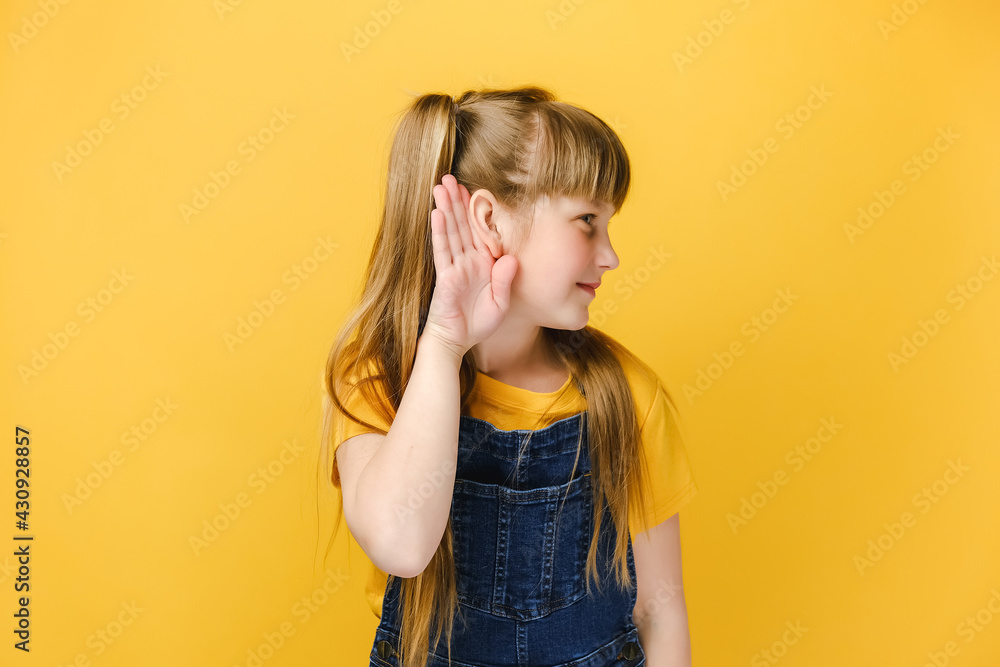 Curious little girl kid likes gossips, wants to overhear secret information, tries to hear conversation, listens attentively keeps hand near ear for eavesdropping, isolated on yellow studio background Curious little girl kid likes gossips, wants to overhear secret information, tries to hear conversation, listens attentively keeps hand near ear for eavesdropping, isolated on yellow studio background