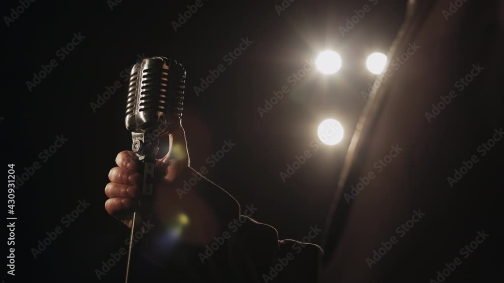 The singer is taking a retro microphone with his hand. The spotlight is shining on him. He's wearing a suit. Close-up shooting. 4K