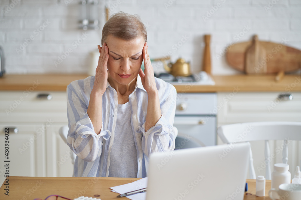 Feeling sick. Stressed mature woman with closed eyes suffering from headache while sitting in kitchen at home, laptop and medicines on wooden table. Healthcare and senior people concept Feeling sick. Stressed mature woman with closed eyes suffering from headache while sitting in kitchen at home, laptop and medicines on wooden table. Healthcare and senior people concept
