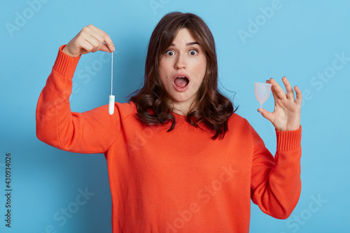 Young attractive brunette female wearing casual clothing holding tampon and menstrual cup in hands, being shocked to know about alternative variant of protection, isolated over blue background.