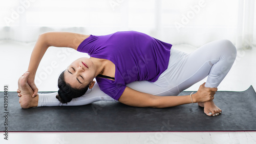 Asian beautiful sportive healthy woman with hair tied wearing purple shirt and white pants, doing exercise, yoga and stretching her body by sitting on mat at home in the morning.