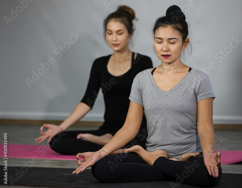 Two Asian beautiful sportive healthy women wearing shirt and black elastic pants, doing exercise, sitting on yoga mat and practicing peacefully meditation and concentration in indoor gym or home.