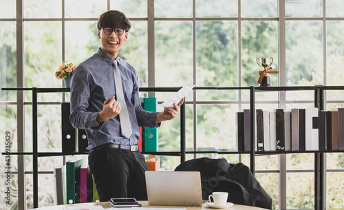 Asian handsome business working man wearing eyeglasses, formal shirt with necktie, large smiling with happiness, raising his arms and doing gesture of winner or success in his project job in office.