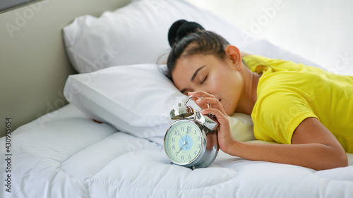 Foto Asian beautiful  lazy woman with hair tied wearing yellow shirt, waking up and lying down on white bed in bedroom at home and touching to snooze time alarm clock in the early morning