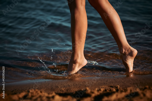 A barefoot girl enters the water to swim. Legs of a slender girl close-up. Rest at a resort near the sea, river. Beach season. Splashes are flying from under your feet © Anton Prokopenko