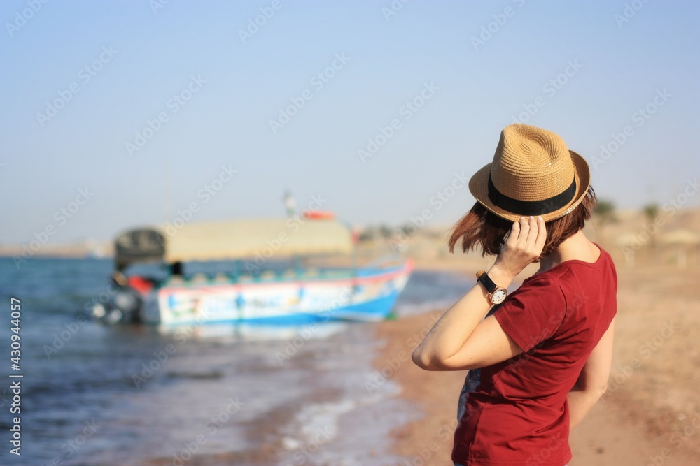 The girl stands on the beach and holds on to her hat with one hand. The girl's face is not visible. The blurry outline of a boat in the background. Sunny day.