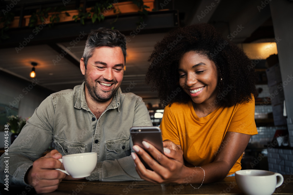 Mixed race couple giggling at pictures on social media holding smartphone in trendy cafe shop Mixed race couple giggling at pictures on social media holding smartphone in trendy cafe shop