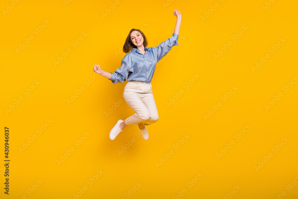 Photo of funny lucky young lady dressed blue outfit jumping high rising fists smiling isolated yellow color background