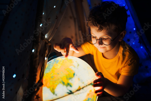 Tableau sur toile A little boy in glasses sits at night in a tent and plays with a globe