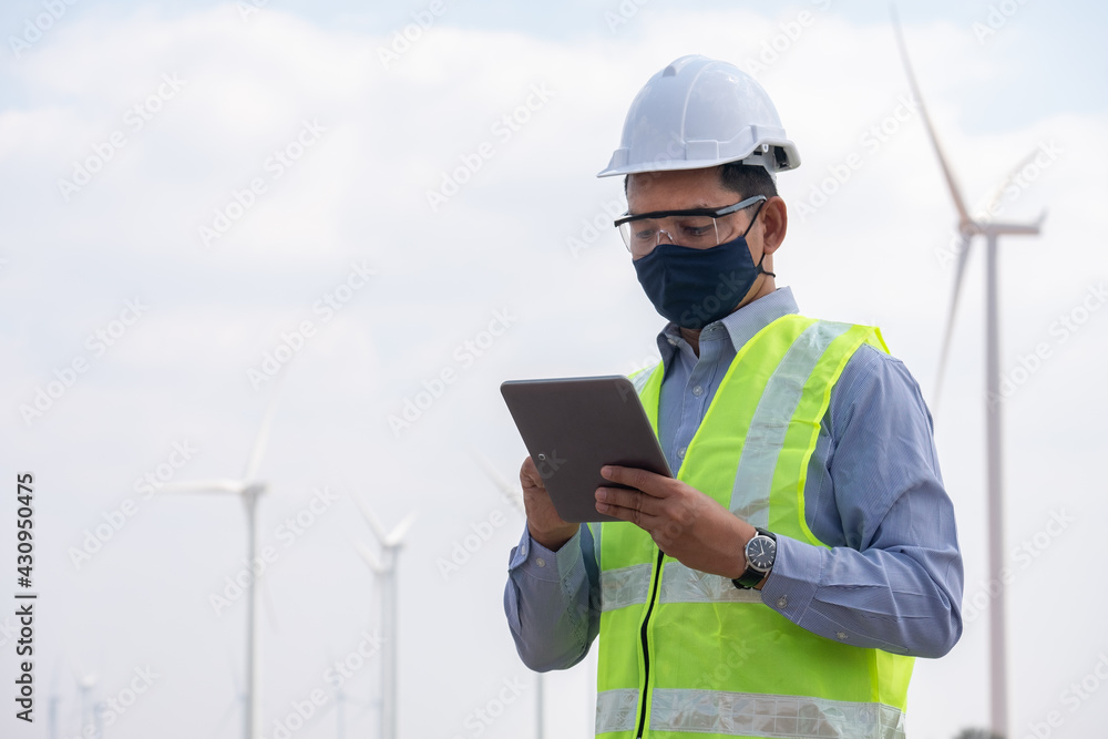 Engineer windmills wearing face mask and hoding tablet with wind turbine in background Engineer windmills wearing face mask and hoding tablet with wind turbine in background