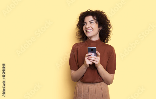 Adorable curly haired woman chatting on mobile posing on a yellow studio wall with free space