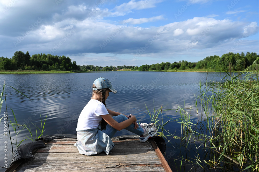 thoughtful young girl white T-shirt, jeans and cap sits wooden pier with her back to the river bank summer sunny weather and enjoys the view river. Time solitude and meditation thoughtful young girl white T-shirt, jeans and cap sits wooden pier with her back to the river bank summer sunny weather and enjoys the view river. Time solitude and meditation