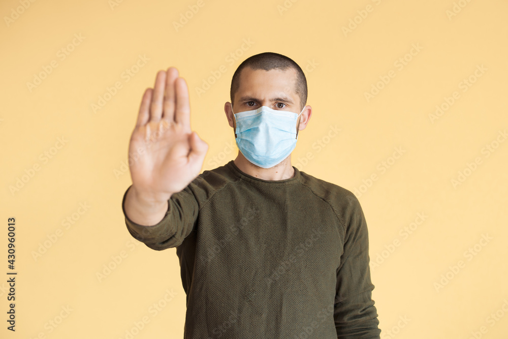 Short haired man is gesturing the stop sign with palm posing on a yellow studio wall wearing a medical mask Short haired man is gesturing the stop sign with palm posing on a yellow studio wall wearing a medical mask