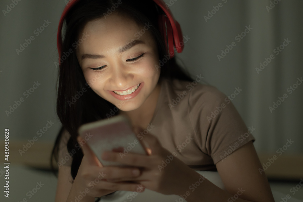 Cute young Asian woman sitting alone with relax and happy emotion on bed at night and using smartphone connect to internet and social media in the dark with light on her face Cute young Asian woman sitting alone with relax and happy emotion on bed at night and using smartphone connect to internet and social media in the dark with light on her face