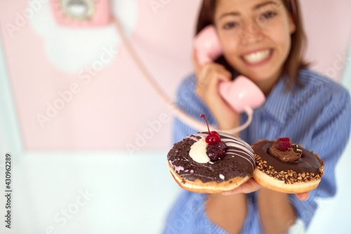 Closeup of a young beautiful girl who is having an interesting conversation on the phone and posing for a photo in a retro looking pastry shop while holding chocolate donuts in her hand