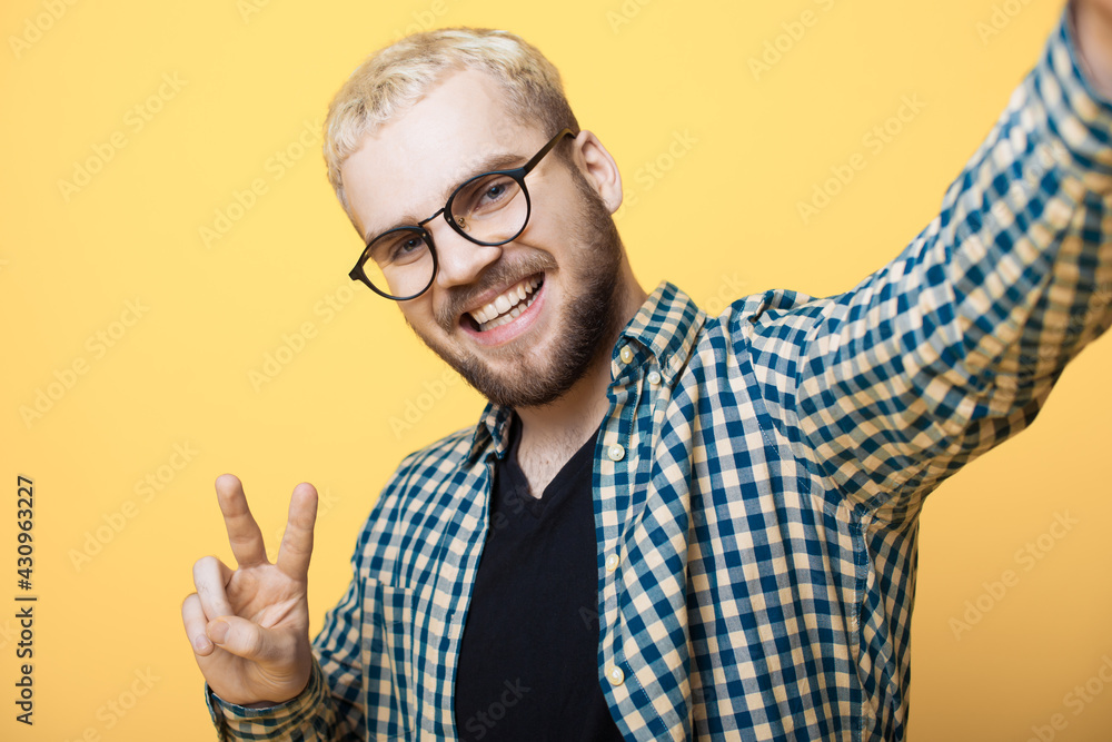 Naklejka premium Caucasian man with beard and blonde hair is making a selfie on a yellow studio wall smiling and gesturing peace sign