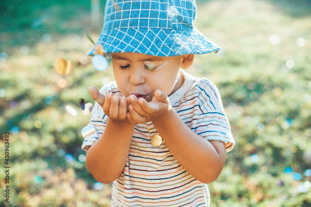 Small caucasian boy is blowing confetti from his palm playing in the garden wearing a blue hat Small caucasian boy is blowing confetti from his palm playing in the garden wearing a blue hat