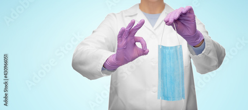 medicine, health protection and healthcare concept - close up of female doctor in white coat and gloves with medical mask showing ok gesture over blue background