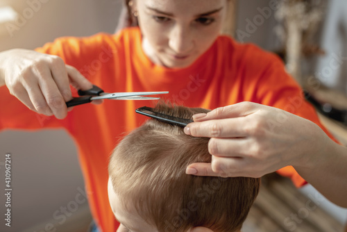 A young woman in bright clothes is cutting a little boy's hair with scissors. Concept of family, care, lifestyle