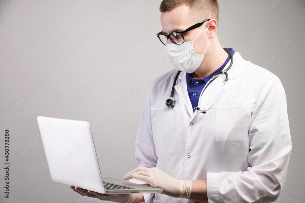 young caucasian doctor in a mask uses his laptop for medical purposes on a gray background. Studio shot young caucasian doctor in a mask uses his laptop for medical purposes on a gray background. Studio shot
