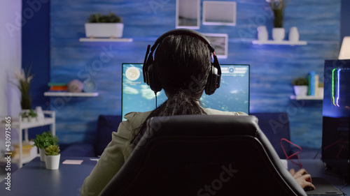Back shot of streamer woman playing on RGB powerful computer shooter video game for tournament, talking with multiple players into headphones. Pro video gamer using professional streaming equipment