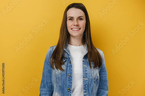 Portrait of good lovely young brunette woman 20s dressed in denim jacket posing while standing and looking at camera isolated over yellow studio background. People, youth, happy feelings concept