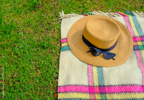 elegant straw female hat with black ribbon, elegant sunglasses on checkered blanket on grass. top view. summer picnic in the park. Copy space