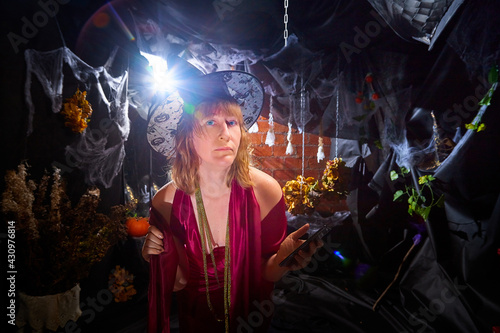 Blonde witch in red dress and black hat in Halloween decoration indoors with cell phone. Woman taking a selfie during holiday carnival
