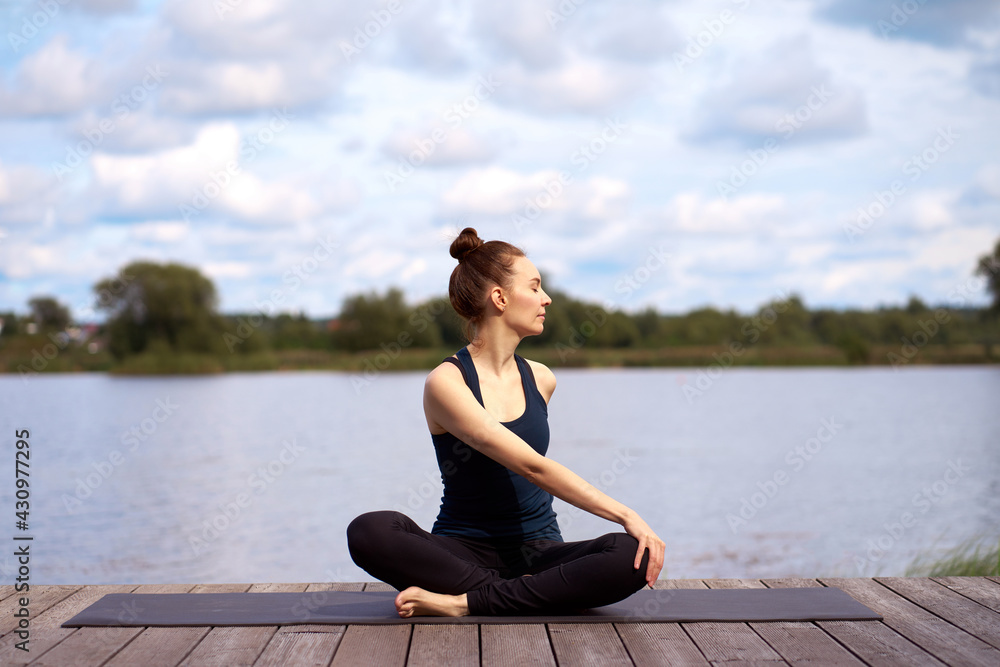Young attractive woman with closed eyes practicing yoga on the wooden pier near lake. Outdoor activities.