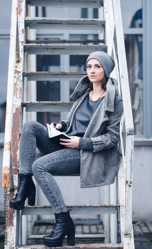 A young girl in a warm gray hat with long hair in a gray sweatshirt and jeans is reading a book while sitting on the metal steps in the industrial zone of the factory A young girl in a warm gray hat with long hair in a gray sweatshirt and jeans is reading a book while sitting on the metal steps in the industrial zone of the factory
