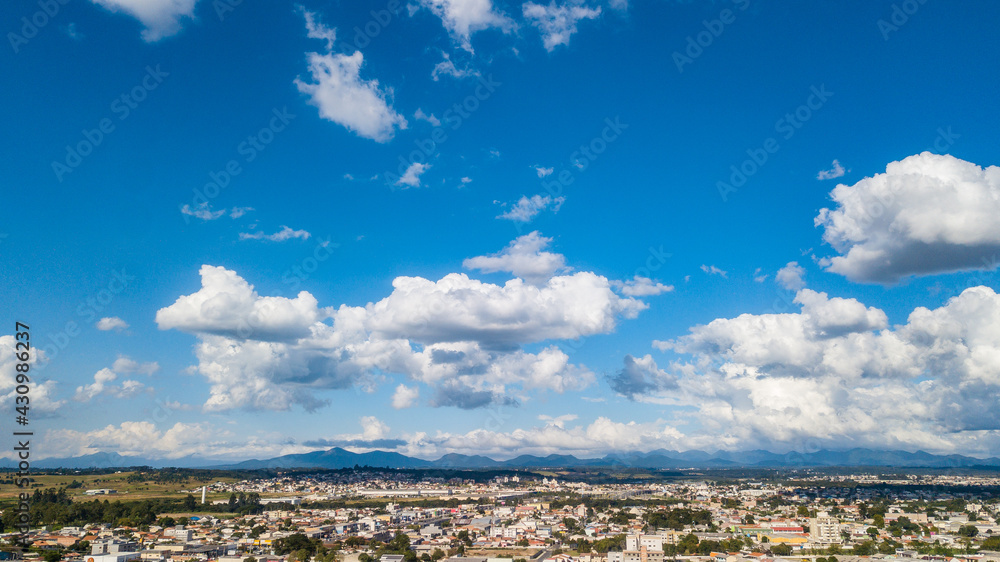 Image of a day with the blue sky and some white clouds. At the base of the image is a city and mountains in the background. 
