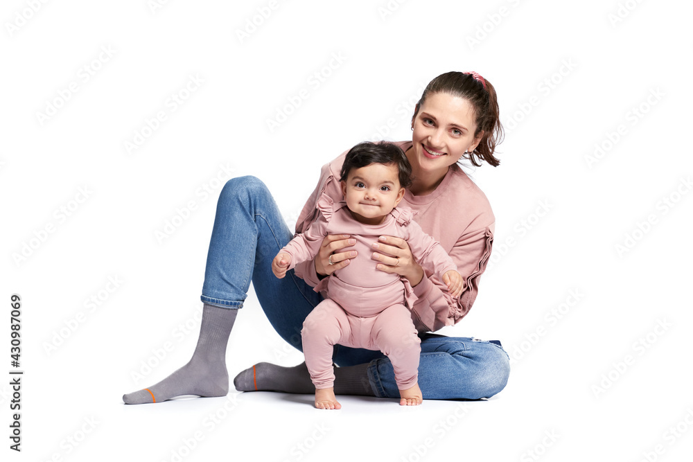 Portrait of cute mother with daughter looking at camera isolated on white studio background. Young attractive woman sitting on floor and helping sweet adorable child to stand, happy childhood concept.