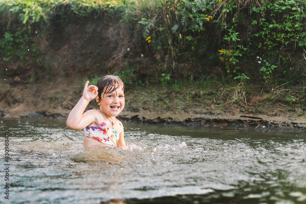 Fototapeta premium Happy summertime, healthy childhood concept. Little girl swimming, splashing, jumping and having fun in a river in summer. Blured green background. Horizontal shot.