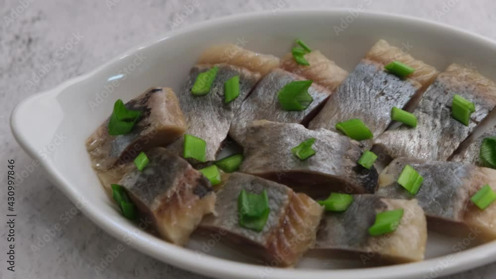 Pieces of herring fillet are sprinkled with green onions on a white plate. Salted sea fish, a traditional cold appetizer. Close-up, top view. Pieces of herring fillet are sprinkled with green onions on a white plate. Salted sea fish, a traditional cold appetizer. Close-up, top view.