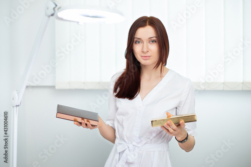 Charming competent female beautician cosmetologist in white uniform posing indoors at modern clinic on camera with cosmetic boxes in hands