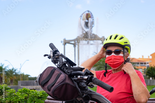 Active senior woman with bicycle in urban public park. She is wearing a protective face mask due to coronavirus and a yellow helmet.Fountain in background
