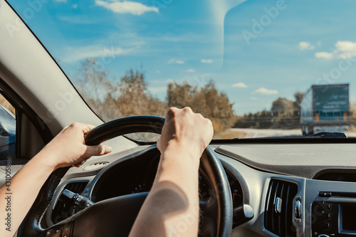 Female hands confidently hold steering wheel of car while driving on suburban highway with traffic. Overtaking truck on road, inside view of dashboard. Woman driving rental car